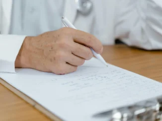 Pessoa idosa escrevendo em um caderno com uma caneta, sentado à mesa de madeira. A imagem transmite cuidado, saúde e envelhecimento ativo.
