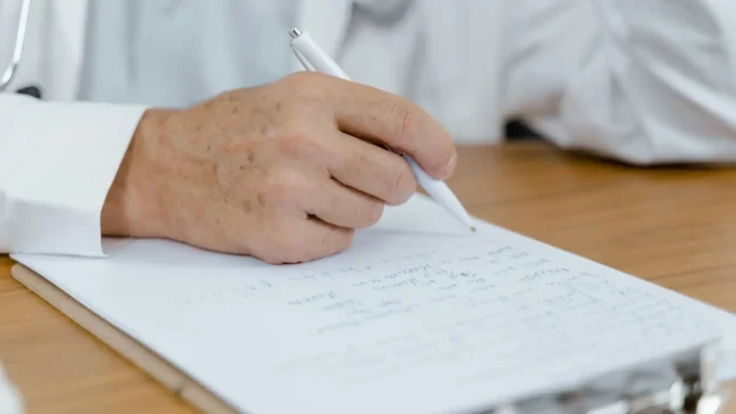 Pessoa idosa escrevendo em um caderno com uma caneta, sentado à mesa de madeira. A imagem transmite cuidado, saúde e envelhecimento ativo.